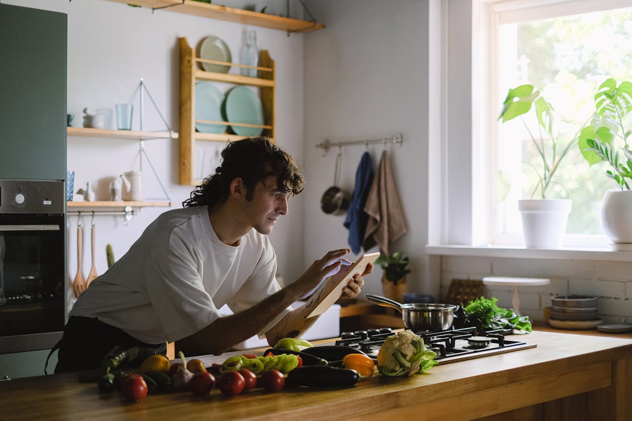 gallery-1 Man preparing food in the kitchen using a tablet for recipe guidance. Vibrant and modern setting.